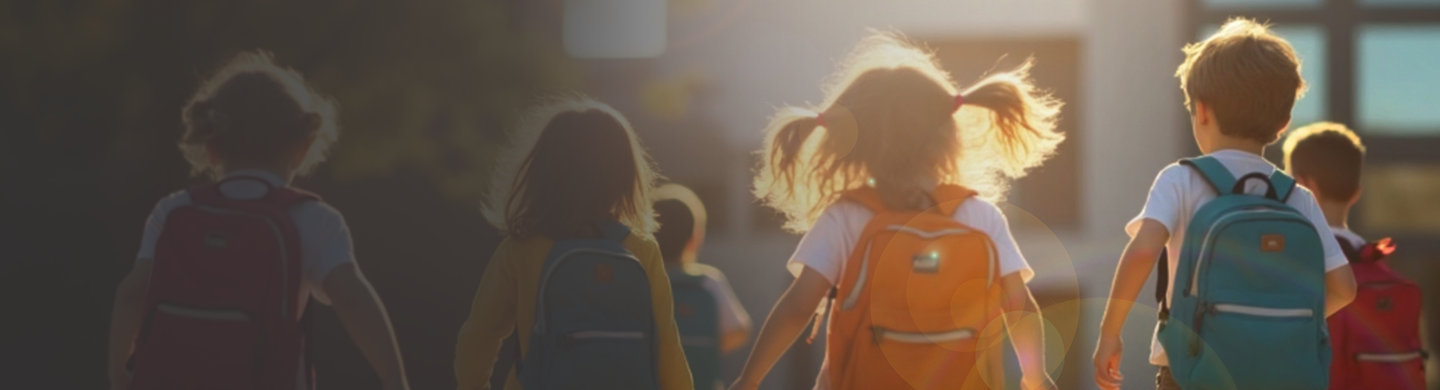 Children wearing backpacks walk toward a school building in warm morning sunlight, viewed from behind, as part of a back-to-school scene.
